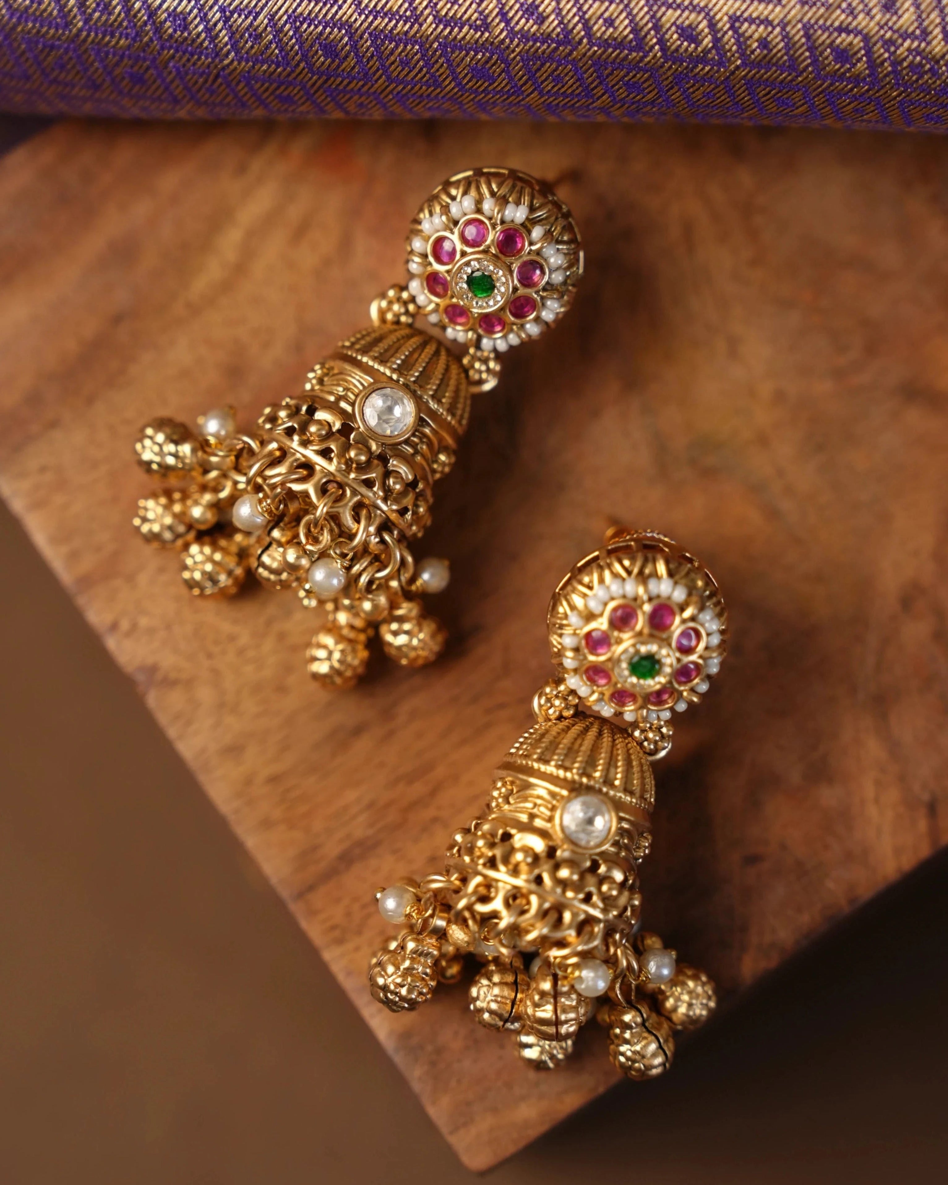 A pair of ornate gold-toned Indian jhumka earrings decorated with red, green, and white stones and small pearl-like beads, displayed on a wooden block with a purple patterned fabric in the background.