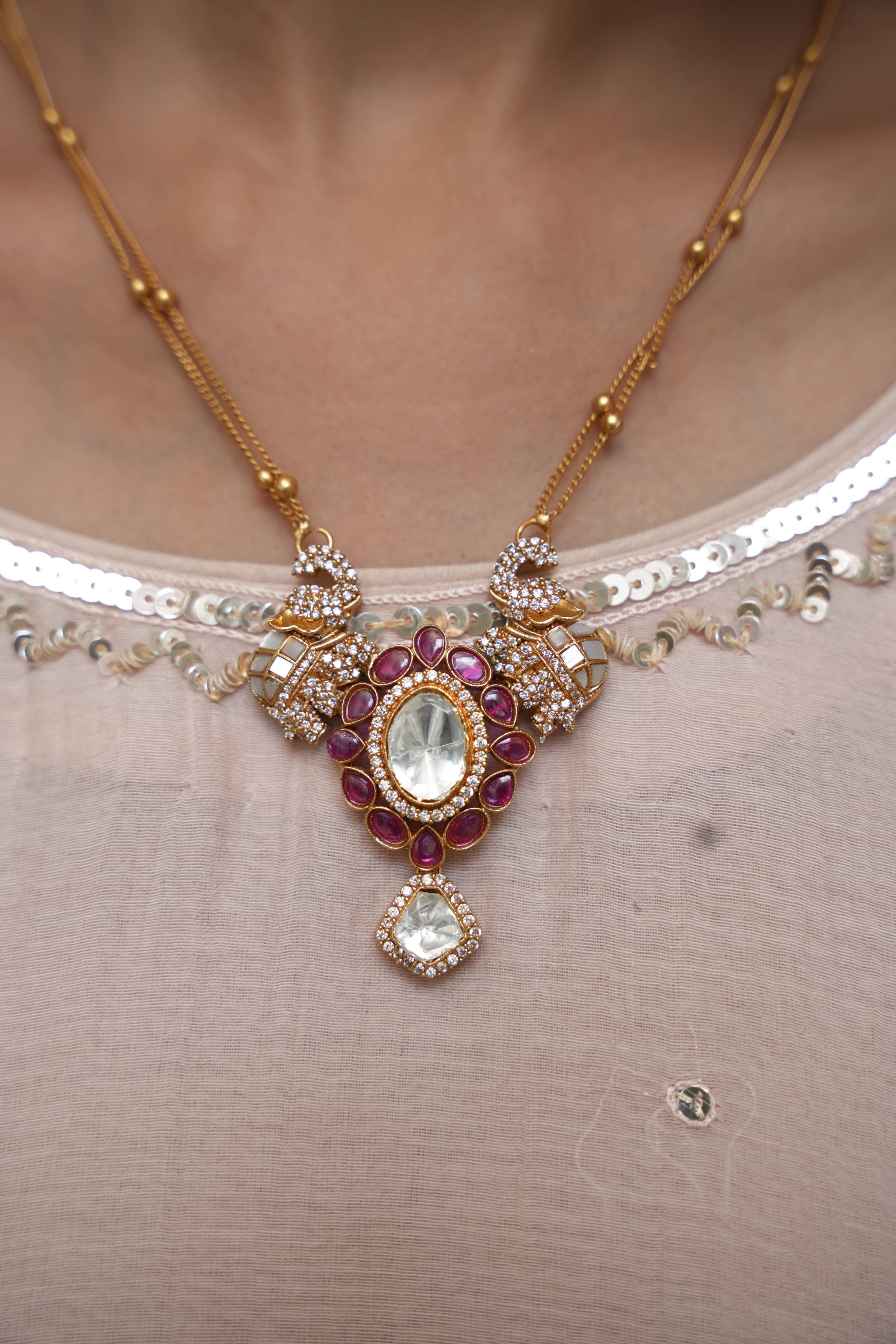 Gold-toned traditional necklace with a detailed pendant featuring red stones and clear crystals, worn on a model's neck over a beige embellished top.