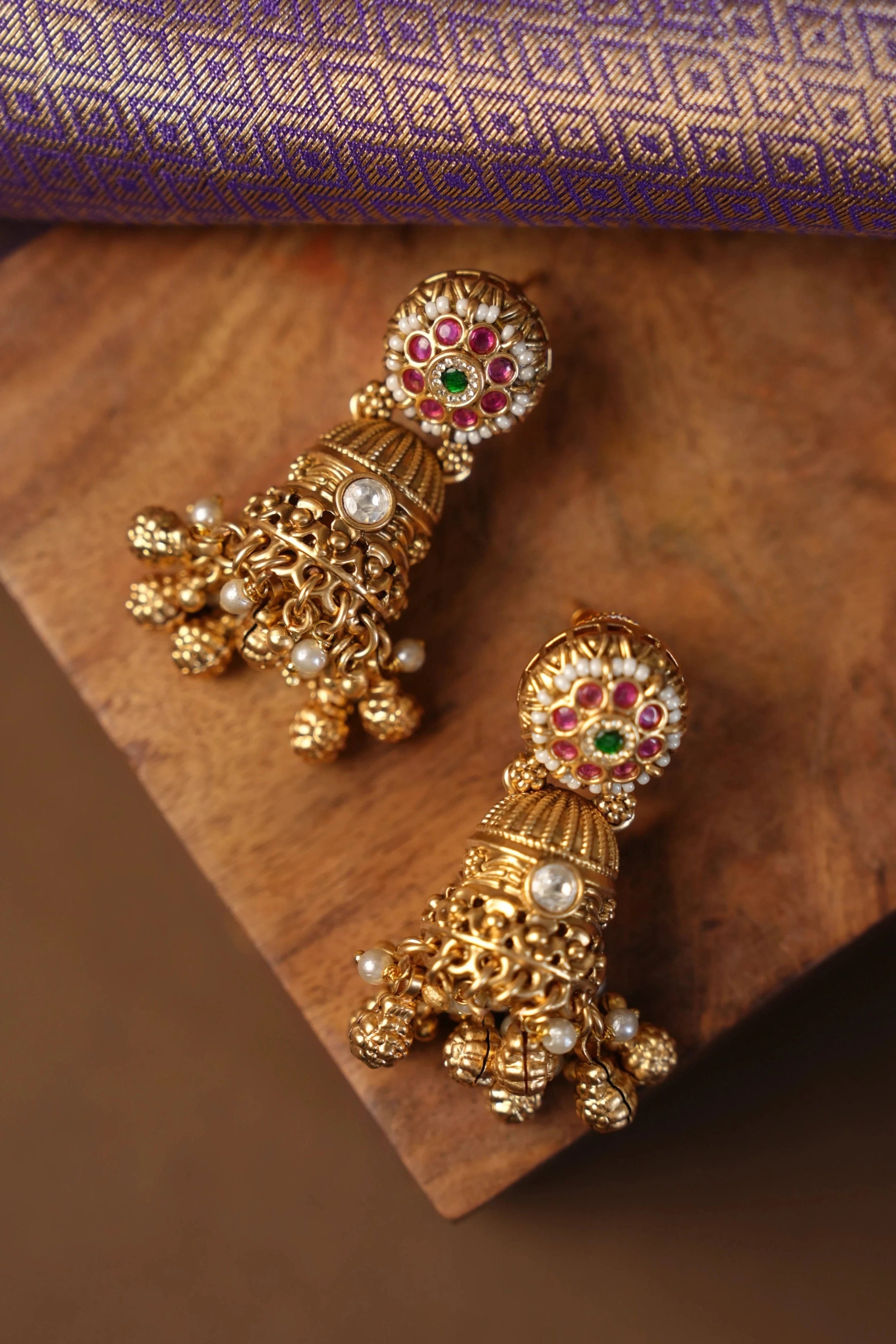 A pair of ornate gold-toned Indian jhumka earrings decorated with red, green, and white stones and small pearl-like beads, displayed on a wooden block with a purple patterned fabric in the background.